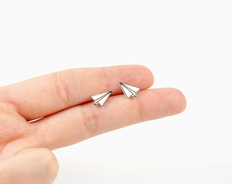 May include: Two small, white paper airplane earrings with black outlines. The earrings are displayed on a light background. The paper airplane design is a classic and whimsical motif.
