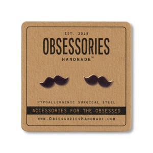 May include: A pair of dark mustache stud earrings on a brown card. The card reads "OBSESSORIES HANDMADE" and "ACCESSORIES FOR THE OBSESSED". The earrings are made of hypoallergenic surgical steel.