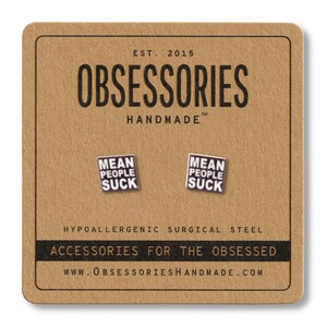 Op de afbeelding: Een paar vierkante oorbellen met de tekst "MEAN PEOPLE SUCK" op een bruine kaart. De kaart toont ook de merknaam "OBSESSORIES HANDMADE" en de tekst "ACCESSORIES FOR THE OBSESSED". De oorbellen zijn gemaakt van hypoallergeen chirurgisch staal.