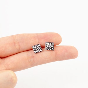Op de afbeelding: Twee vierkante zwart-witte oorbellen. Elke oorbel heeft de tekst "MEAN PEOPLE SUCK" in een vetgedrukte, schreefloze lettertype. De oorbellen zijn klein en lijken van glanzend materiaal te zijn. Ze worden gepresenteerd tegen een lichte achtergrond.