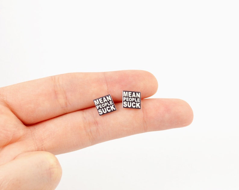 Op de afbeelding: Twee vierkante zwart-witte oorbellen. Elke oorbel heeft de tekst "MEAN PEOPLE SUCK" in een vetgedrukte, schreefloze lettertype. De oorbellen zijn klein en lijken van glanzend materiaal te zijn. Ze worden gepresenteerd tegen een lichte achtergrond.