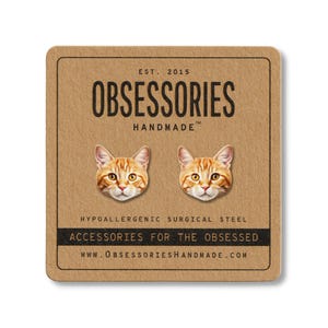 May include: A pair of orange tabby cat face earrings on a brown card. The card reads "OBSESSORIES HANDMADE" and "ACCESSORIES FOR THE OBSESSED". The earrings are made of hypoallergenic surgical steel.