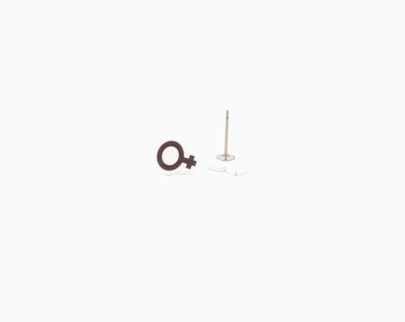 May include: A pair of stud earrings featuring the female gender symbol. The earrings have a dark brown outline with a white center. One earring is displayed upright, showing the post and backing.