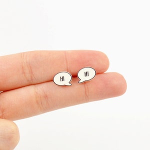 May include: A pair of small, speech bubble-shaped earrings. Each earring is white with a black outline and features the word "HI" in black. The earrings are held against a white background.