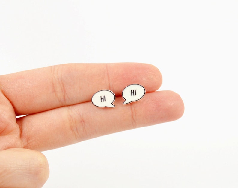 May include: A pair of small, speech bubble-shaped earrings. Each earring is white with a black outline and features the word "HI" in black. The earrings are held against a white background.