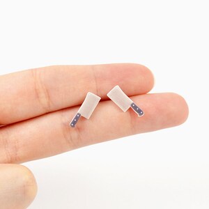 May include: A pair of miniature cleaver earrings. The earrings have a silver-colored blade and a handle with three small, dark blue dots. The earrings are small and delicate, perfect for adding a touch of whimsy to any outfit.