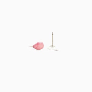 May include: A single pink petal and a silver earring post with a white backing are set against a white background. The petal has a delicate, translucent appearance with a gradient of pink hues.