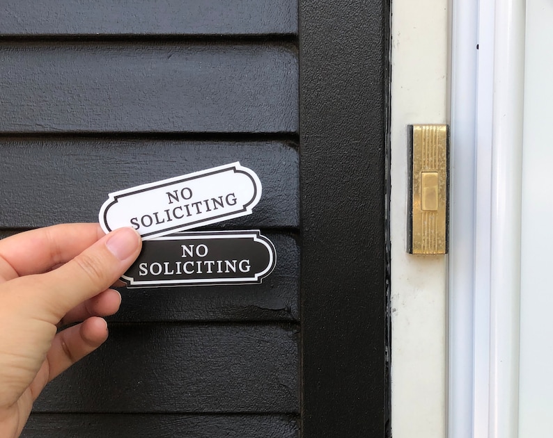 Puede incluir: Dos letreros en blanco y negro con el texto "No Soliciting" en una puerta negra. Los letreros est&aacute;n pegados a la puerta con adhesivo.