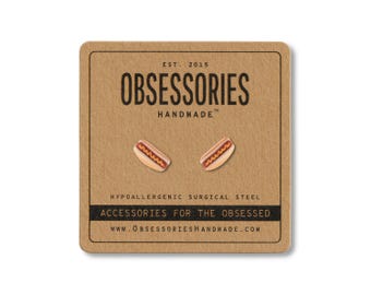 May include: A pair of hot dog-shaped earrings on a brown card. The card has the text "OBSESSORIES HANDMADE" and "ACCESSORIES FOR THE OBSESSED". The earrings are made of hypoallergenic surgical steel.
