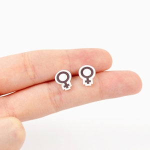 May include: Two small, white earrings with a black female symbol design. The earrings are held in a person's hand against a white background. The earrings are small and simple, with a minimalist design.