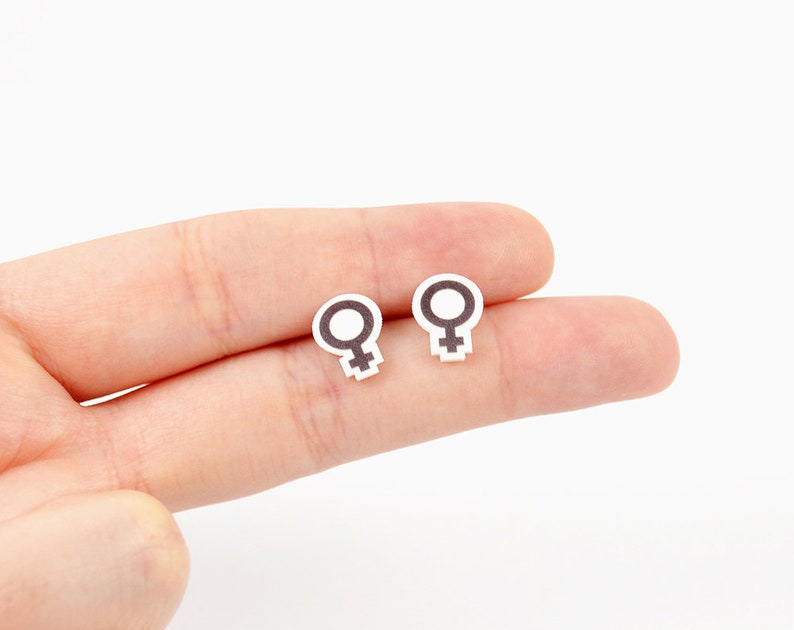 May include: Two small, white earrings with a black female symbol design. The earrings are held in a person's hand against a white background. The earrings are small and simple, with a minimalist design.
