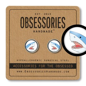 May include: A pair of round, silver-toned stud earrings featuring a cartoon shark design with a blue and white color scheme. The earrings are displayed on a brown card with the text "OBSESSORIES HANDMADE" and "ACCESSORIES FOR THE OBSESSED".