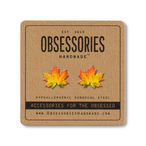 May include: A pair of maple leaf stud earrings in shades of red, orange, and yellow, set against a brown card. The card reads "OBSESSORIES HANDMADE" and "ACCESSORIES FOR THE OBSESSED". The earrings are made of hypoallergenic surgical steel.