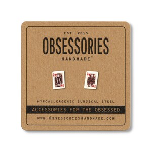 Puede incluir: Pendientes con forma de carta de juego, presentados en una tarjeta marrón. La tarjeta presenta el texto "OBSESSORIES HANDMADE" y "ACCESSORIES FOR THE OBSESSED". Los pendientes son de acero quirúrgico hipoalergénico.