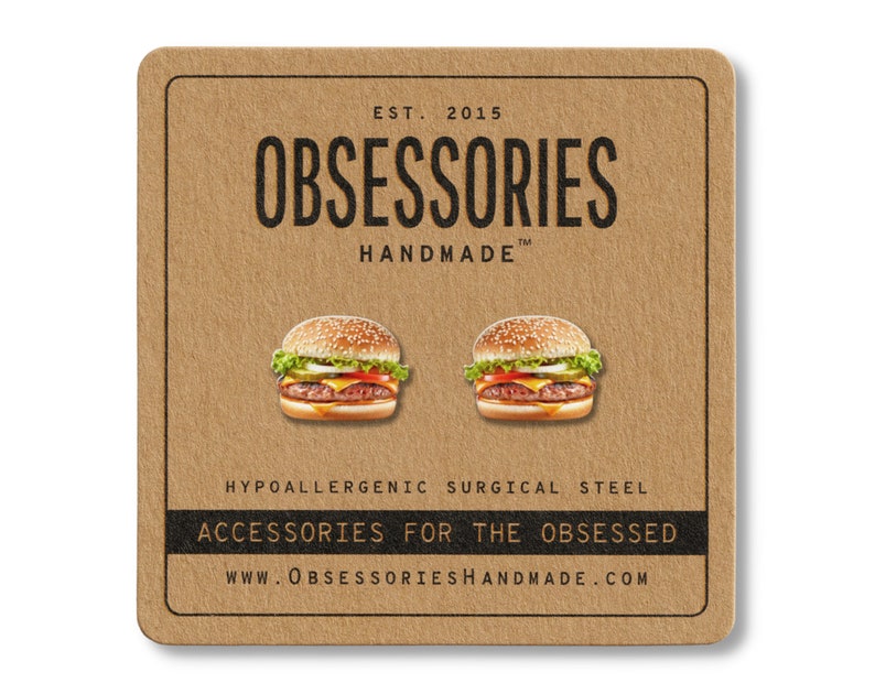 May include: A pair of burger-shaped earrings on a brown card. The earrings are detailed with sesame seed buns, a beef patty, cheese, lettuce, and tomato. The card reads "OBSESSORIES HANDMADE" and "ACCESSORIES FOR THE OBSESSED."