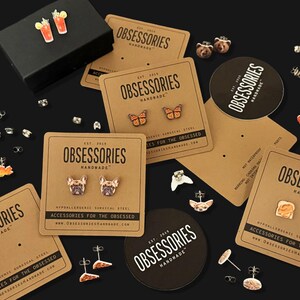 May include: An assortment of stud earrings on display, including butterfly, dog, and food-themed designs. The earrings are presented on branded cards and a black surface. A black box with cocktail earrings is also present.