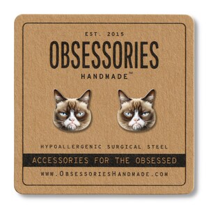 May include: A pair of cat face stud earrings on a brown card. The earrings feature the face of a grumpy cat. The card reads "OBSESSORIES HANDMADE" and "ACCESSORIES FOR THE OBSESSED". The website address is also included.