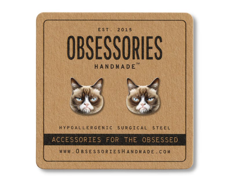 May include: A pair of cat face stud earrings on a brown card. The earrings feature the face of a grumpy cat. The card reads "OBSESSORIES HANDMADE" and "ACCESSORIES FOR THE OBSESSED". The website address is also included.