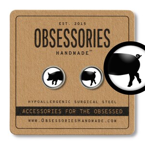 Puede incluir: Un par de pendientes de botón plateados con una silueta de cerdo negro sobre un fondo blanco. Los pendientes se muestran en una tarjeta marrón con el texto "OBSESSORIES HANDMADE" y "ACCESSORIES FOR THE OBSESSED".