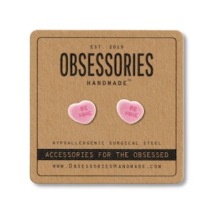 May include: A pair of pink heart-shaped stud earrings with the words "BE MINE" printed on them. The earrings are displayed on a brown card with the text "OBSESSORIES HANDMADE" and "ACCESSORIES FOR THE OBSESSED".