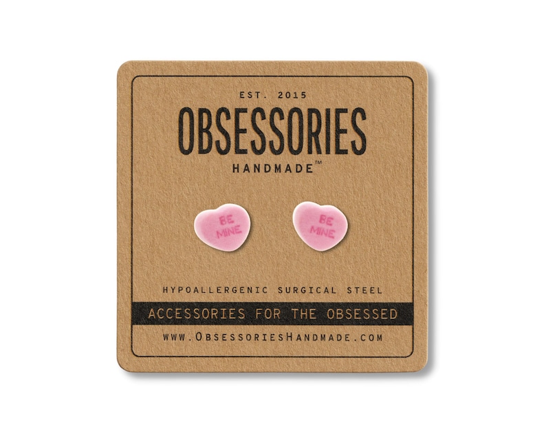 May include: A pair of pink heart-shaped stud earrings with the words "BE MINE" printed on them. The earrings are displayed on a brown card with the text "OBSESSORIES HANDMADE" and "ACCESSORIES FOR THE OBSESSED".