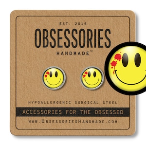 Puede incluir: Un par de pendientes amarillos con forma de cara sonriente con salpicaduras de sangre roja en un lado. Los pendientes están hechos de acero quirúrgico hipoalergénico. El texto en la tarjeta dice "EST. 2015 OBSESSORIES HANDMADE TM HYPOALLERGENIC SURGICAL STEEL ACCESSORIES FOR THE OBSESSED WWW.OBSESSORIESHANDMADE.COM"