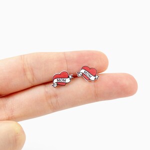 K&ouml;nnte beinhalten: Ein Paar kleine Ohrstecker mit einem roten Herz-Design und einem Banner mit der Aufschrift "MOM". Die Ohrringe haben eine klassische Tattoo-&Auml;sthetik mit wei&szlig;em Rand und silberfarbener R&uuml;ckseite.