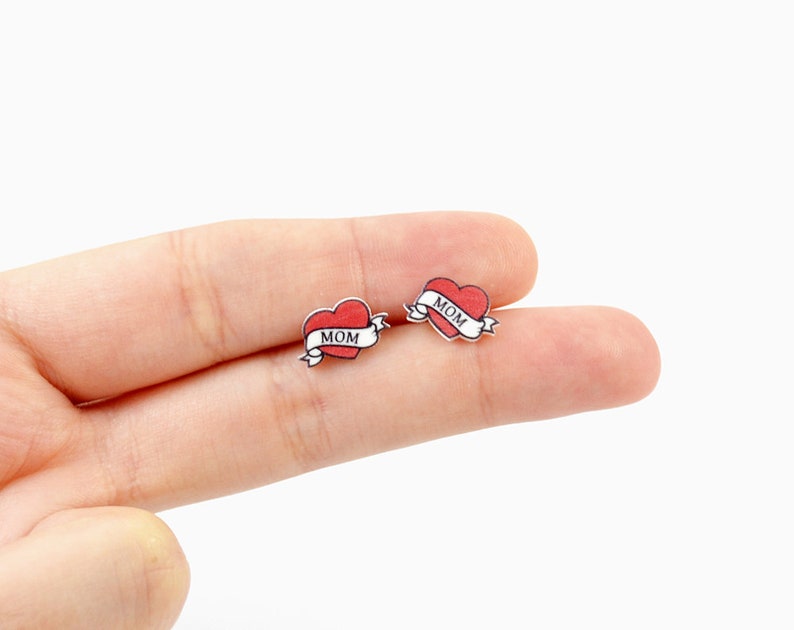 K&ouml;nnte beinhalten: Ein Paar kleine Ohrstecker mit einem roten Herz-Design und einem Banner mit der Aufschrift "MOM". Die Ohrringe haben eine klassische Tattoo-&Auml;sthetik mit wei&szlig;em Rand und silberfarbener R&uuml;ckseite.