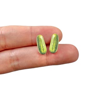 May include: A pair of novelty earrings shaped like miniature pickles. The earrings are green with yellow highlights, mimicking the appearance of a real pickle. The earrings are small and appear to be made of a lightweight material.