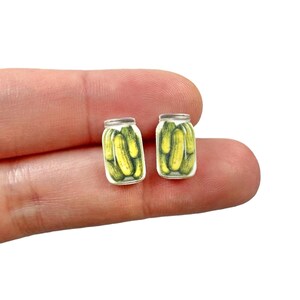 May include: A pair of small, novelty earrings featuring miniature jars of pickles. Each earring depicts a clear jar with a silver lid, filled with bright yellow-green pickles. The earrings are set against a neutral background.