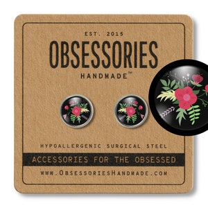 May include: Two round stud earrings with a floral design on a black background. The earrings are presented on a brown card with the text "OBSESSORIES HANDMADE" and "ACCESSORIES FOR THE OBSESSED". The earrings are made of hypoallergenic surgical steel.