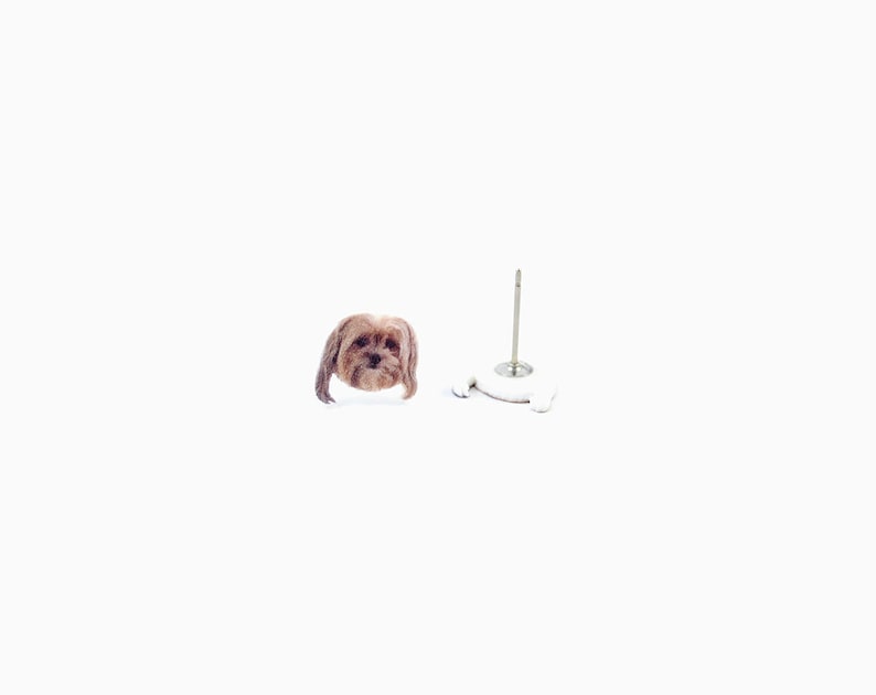 May include: A pair of dog-shaped stud earrings. One earring shows the dog's face, and the other is a white earring post with a small, dog-shaped base. The dog's fur is brown.