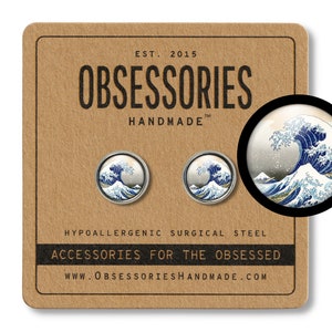 Puede incluir: Un par de pendientes de botón de plata con una reproducción de la famosa estampa en madera "La Gran Ola de Kanagawa" del artista japonés Hokusai. Los pendientes son hipoalergénicos y están hechos de acero quirúrgico. El texto "OBSESSORIES HANDMADE" está impreso en el fondo de la tarjeta marrón.