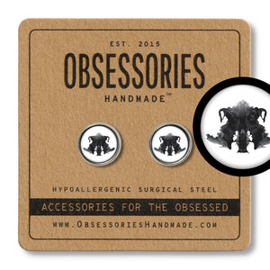 Op de afbeelding: Een paar zilveren oorbel studs met een zwart-witte inktvlek ontwerp. De oorbellen zijn hypoallergeen en gemaakt van chirurgisch staal. De verpakking toont de tekst "OBSESSORIES HANDMADE" en "ACCESSORIES FOR THE OBSESSED".