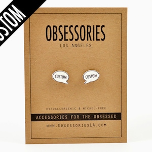 以下が含まれることがあります： 「OBSESSORIES LOS ANGELES」というテキストが書かれた茶色のカードと、「CUSTOM」というテキストが書かれた白い吹き出し型のピンが2つあります。カードには、「HYPOALLERGENIC & NICKEL-FREE ACCESSORIES FOR THE OBSESSED WWW.OBSESSORIESLA.COM」というテキストも書かれています。