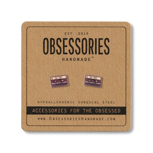 以下が含まれることがあります： 茶色のカードにカセットテープ型のスタッドピアス。カードには「OBSESSORIES HANDMADE」と「ACCESSORIES FOR THE OBSESSED」と書かれています。ピアスは紫と白です。