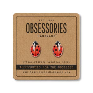 May include: A pair of ladybug stud earrings on a brown card. The earrings are red with black spots and black heads. The card reads "OBSESSORIES HANDMADE" and "ACCESSORIES FOR THE OBSESSED". The earrings are made of hypoallergenic surgical steel.