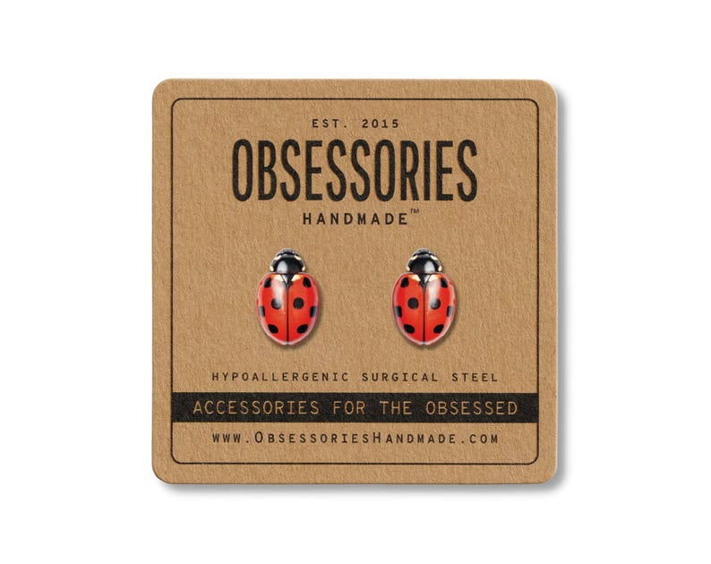 May include: A pair of ladybug stud earrings on a brown card. The earrings are red with black spots and black heads. The card reads "OBSESSORIES HANDMADE" and "ACCESSORIES FOR THE OBSESSED". The earrings are made of hypoallergenic surgical steel.