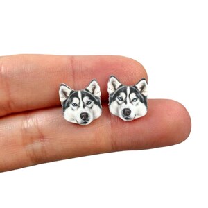 May include: A pair of stud earrings featuring a detailed depiction of a Husky dog's face. The earrings showcase the dog's distinctive black and white markings, with blue eyes, and are held in a person's hand.