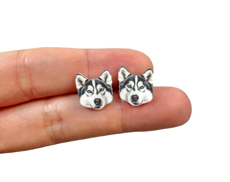 May include: A pair of stud earrings featuring a detailed depiction of a Husky dog's face. The earrings showcase the dog's distinctive black and white markings, with blue eyes, and are held in a person's hand.