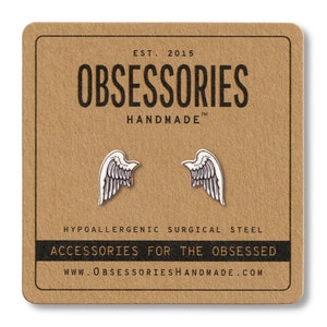 Puede incluir: Pendientes plateados con forma de ala sobre una tarjeta marr&oacute;n. La tarjeta tiene el texto "OBSESSORIES HANDMADE" y "ACCESSORIES FOR THE OBSESSED". Los pendientes son de acero quir&uacute;rgico hipoalerg&eacute;nico.