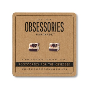 May include: A pair of Polaroid camera stud earrings on a brown card. The card has the text "OBSESSORIES HANDMADE" and "ACCESSORIES FOR THE OBSESSED". The earrings are white, black, and red.