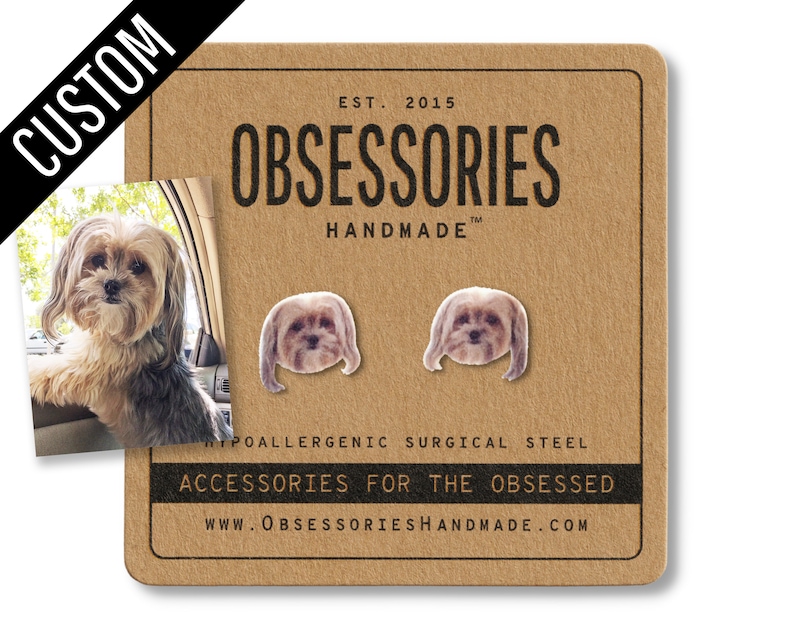 May include: A pair of dog-shaped stud earrings on a brown card. The earrings are small, with a detailed image of a dog's face. The card has the text "OBSESSORIES HANDMADE" and "ACCESSORIES FOR THE OBSESSED". The earrings are made of hypoallergenic surgical steel.