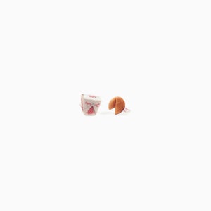 May include: A fortune cookie with a visible fortune and a Chinese takeout box with red text and a tree design. The fortune cookie is brown and open, revealing a white slip of paper. The takeout box is white with red accents.