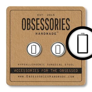 May include: Two silver-colored battery icon pins on a brown background. The text "OBSESSORIES HANDMADE" is printed on the background. The text "HYPOALLERGENIC SURGICAL STEEL ACCESSORIES FOR THE OBSESSED WWW.OBSESSORIESHANDMADE.COM" is printed on the background.