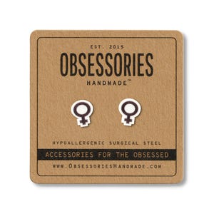 May include: A pair of white and black female symbol stud earrings on a brown card. The card reads "OBSESSORIES HANDMADE" and "ACCESSORIES FOR THE OBSESSED". The earrings are made of hypoallergenic surgical steel.