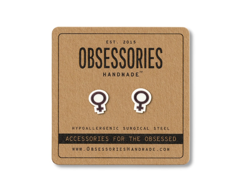 May include: A pair of white and black female symbol stud earrings on a brown card. The card reads "OBSESSORIES HANDMADE" and "ACCESSORIES FOR THE OBSESSED". The earrings are made of hypoallergenic surgical steel.