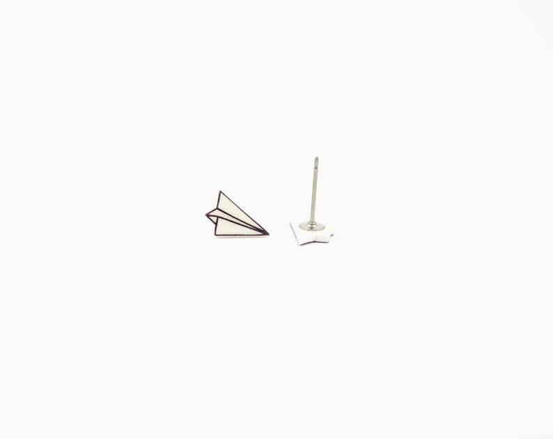 May include: A pair of paper airplane stud earrings. The earrings feature a white paper airplane design with a dark outline. One earring is a complete paper airplane, and the other is the post and backing.