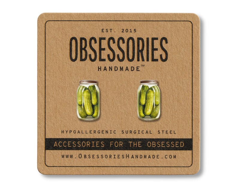 May include: A pair of pickle jar earrings on a brown card. The earrings are miniature jars filled with green pickles. The card has the text "OBSESSORIES HANDMADE" and "ACCESSORIES FOR THE OBSESSED". The earrings are made of hypoallergenic surgical steel.