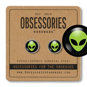 May include: A pair of hypoallergenic surgical steel earrings featuring a green alien head design. The earrings are packaged on a brown card with the text "OBSESSORIES HANDMADE" and "ACCESSORIES FOR THE OBSESSED".